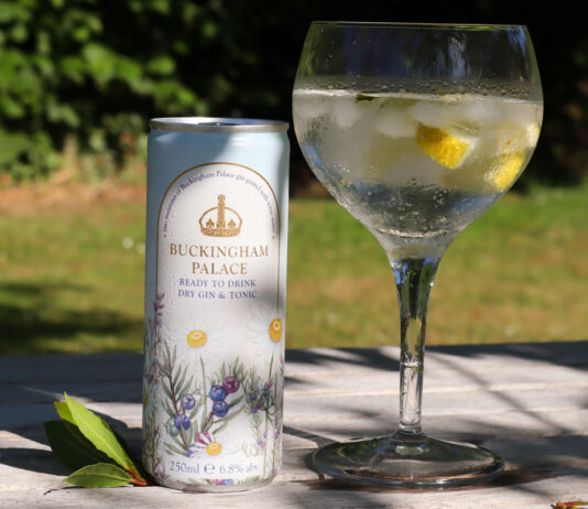 Buckingham Palace enters the RTD cocktail market with new Royal Collection Gin & Tonic cans A can of Buckingham Palace Ready to Drink Dry Gin & Tonic stands next to a balloon glass with the serve inside with lemon peel and ice. The pair sit on a wooden table with greenery in the background.