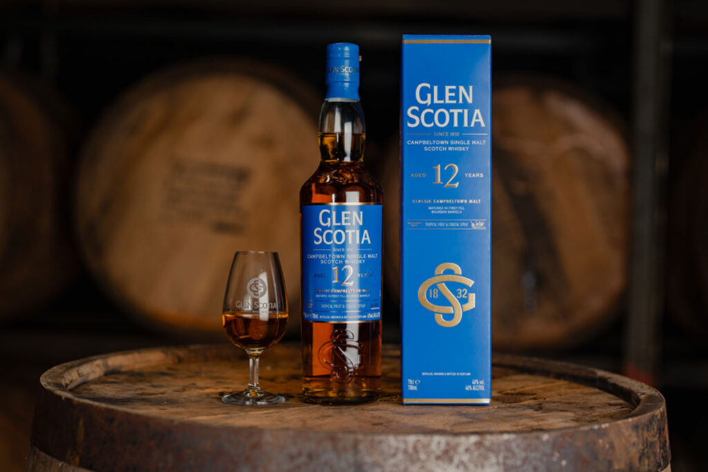 A bottle of Glen Scotia 12 Year Old stands next to its outer box packaging with a dram of the whisky inside a nosing glass next to it. All three stand on top of a barrel.