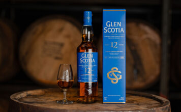A bottle of Glen Scotia 12 Year Old stands next to its outer box packaging with a dram of the whisky inside a nosing glass next to it. All three stand on top of a barrel.