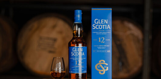 A bottle of Glen Scotia 12 Year Old stands next to its outer box packaging with a dram of the whisky inside a nosing glass next to it. All three stand on top of a barrel.
