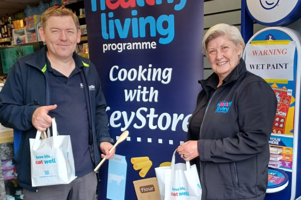 A member of the KeyStore team and a member of the Healthy Living Programme team stand inside a KeyStore shop holding bags as part of a recipe meal initiative with the KeyStore estate.