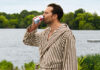 Hollywood actor Jamie Dornan stands in a dressing gown drinking from a can of Diet Coke with a lake behind him.