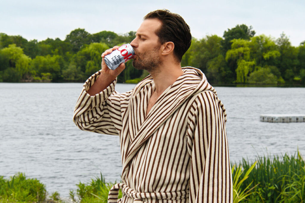 Hollywood actor Jamie Dornan stands in a dressing gown drinking from a can of Diet Coke with a lake behind him.