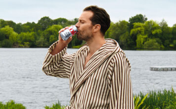 Hollywood actor Jamie Dornan stands in a dressing gown drinking from a can of Diet Coke with a lake behind him.