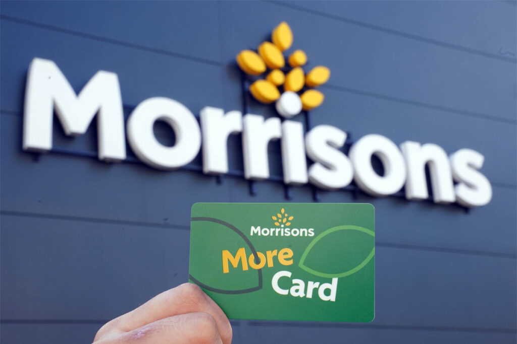A person holds up a Morrisons More card against a Morrisons sign which is on a building with black panelling.