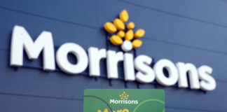 A person holds up a Morrisons More card against a Morrisons sign which is on a building with black panelling.