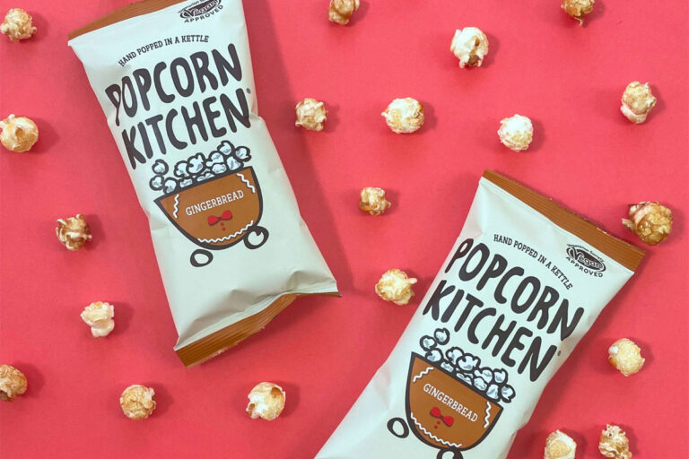 Packets of Popcorn Kitchen Gingerbread Popcorn lie against a red background with pieces of the popcorn dotted around them.