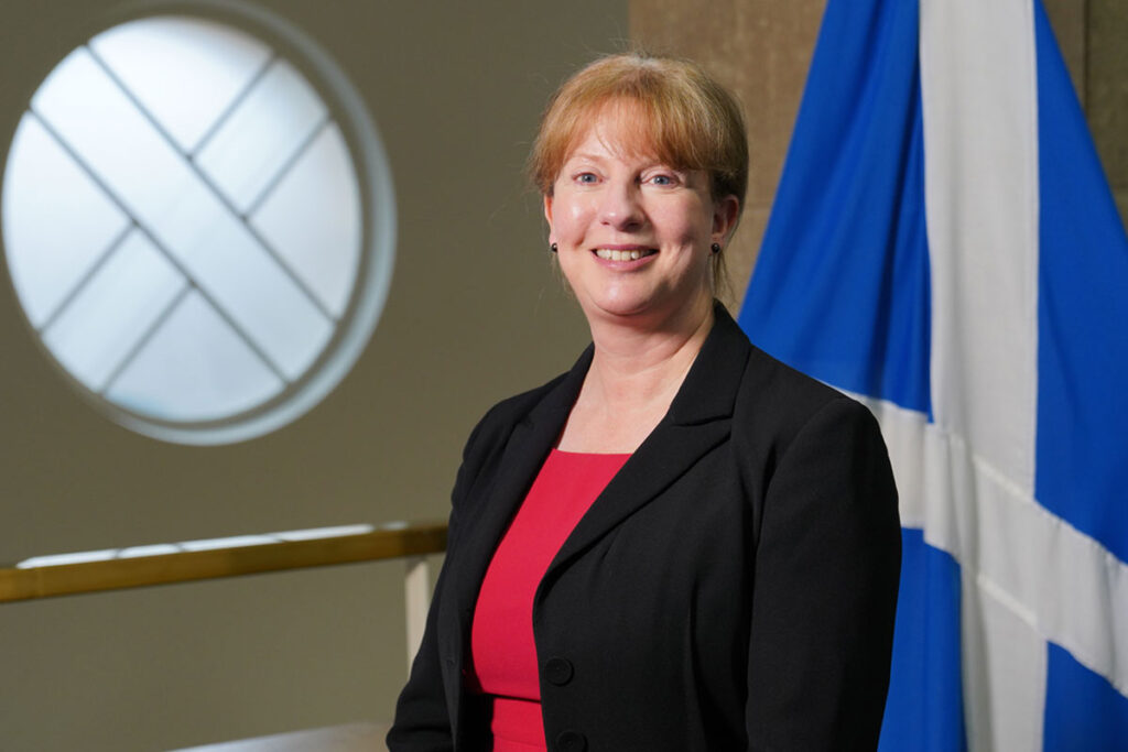 Scottish finance secretary Shona Robison stands in Parliament with the Scotland flag behind her.