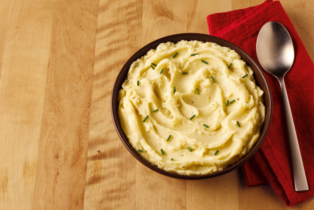 A bowl of mashed potato sits on a table with a red napkin next to it and a spoon on top of the napkin.