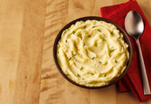 A bowl of mashed potato sits on a table with a red napkin next to it and a spoon on top of the napkin.