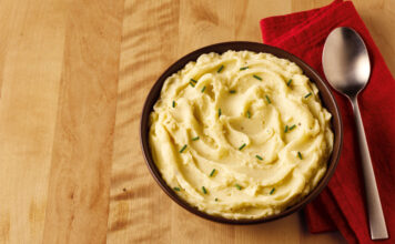 A bowl of mashed potato sits on a table with a red napkin next to it and a spoon on top of the napkin.