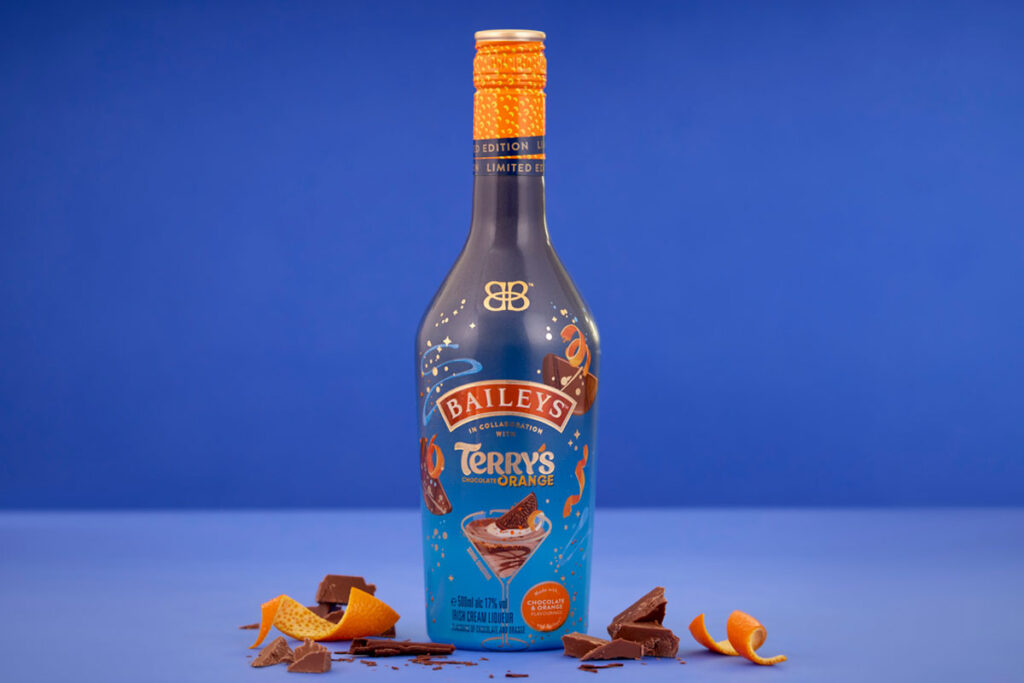 A bottle of Baileys Terry's Chocolate Orange limited edition flavour stands in front of a blue background with orange peel and pieces of chocolate around the bottle.