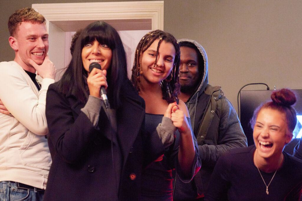 TV host Claudia Winkleman stands with a group of young people holding a microphone in a recording studio.