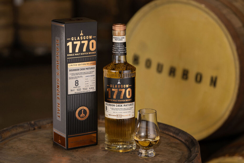 A bottle of Glasgow 1770 Bourbon Cask Matured Single Malt stands on whisky barrel with the outer packaging next to it and a nosing glass with the whisky inside on its other side. There is also a barrel in the background labelled as Bourbon.