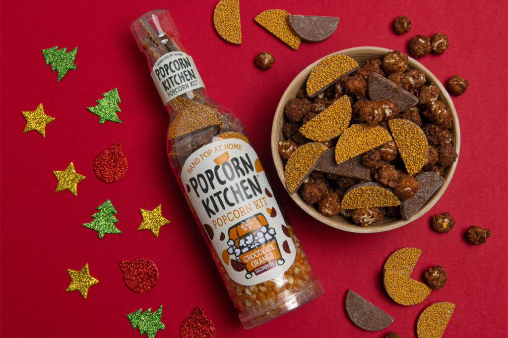 A pack of Popcorn Kitchen Popcorn Kit Chocolate Orange lies on a red surface with a bowl of the popcorn ready-made next to it with Christmas decorations around the bottle.