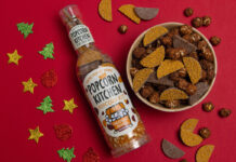A pack of Popcorn Kitchen Popcorn Kit Chocolate Orange lies on a red surface with a bowl of the popcorn ready-made next to it with Christmas decorations around the bottle.