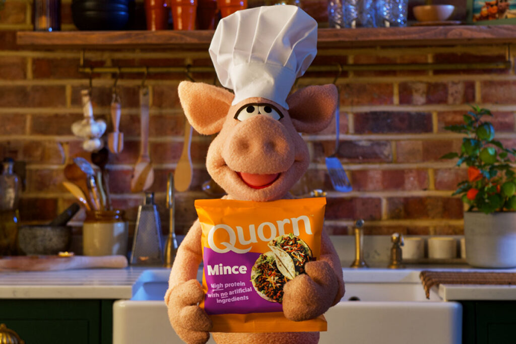 A puppet pig wearing a chef hat stands in a kitchen holding a bag of Quorn Mince.