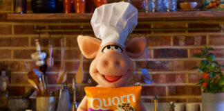 A puppet pig wearing a chef hat stands in a kitchen holding a bag of Quorn Mince.