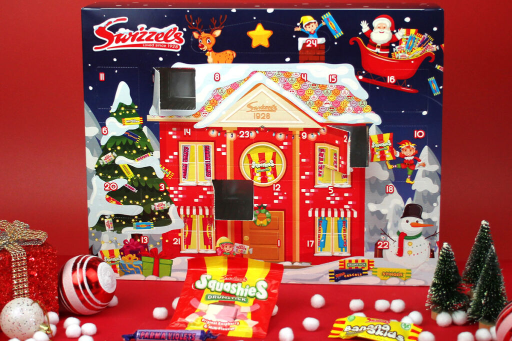 The Swizzels Advent Calendar for 2025 stands against a red background with packets of Swizzels sweets lying in front of the calendar. Doors one the calendar have also been opened.
