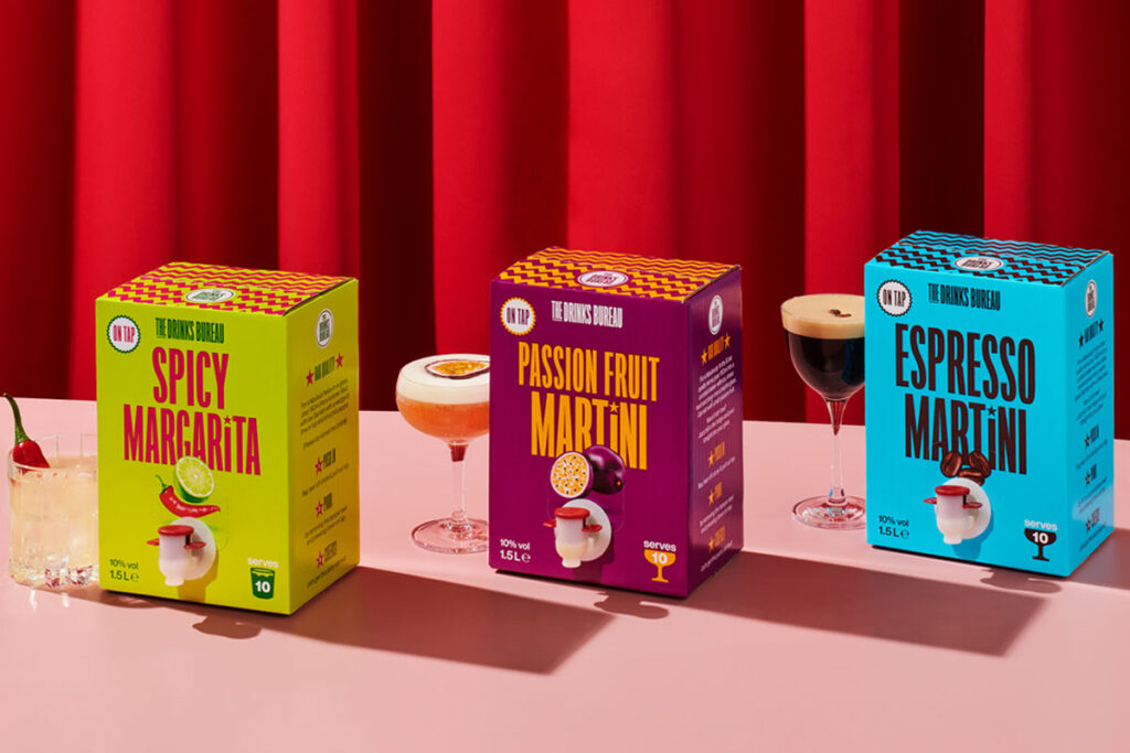 The Drinks Bureau Boxed Cocktails including Spicy Margarita, Passion Fruit Martini and Espresso Martini with the cocktails served in glasses next to each of the boxes with a red curtain in the background.