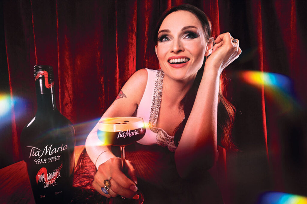 Sophie Ellis Bextor sits at a table holding an Espresso Martini cocktail in a Tia Maria branded glass. There is also a bottle of Tia Maria on the table.