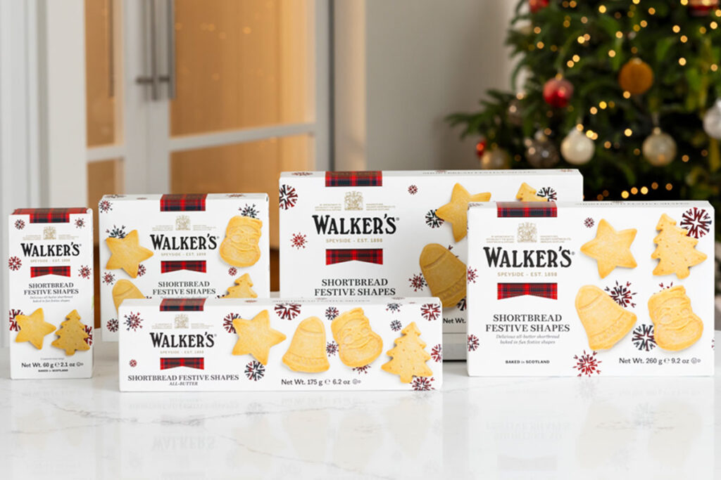 Packs of Walker's Shortbread 2025 Christmas selection stand upright on a white marble table with a Christmas tree in the background.