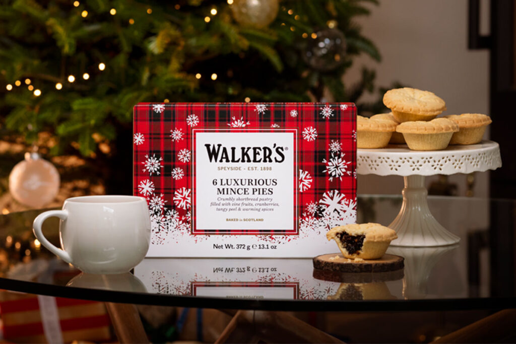 A pack of Walker's Luxurious Mince Pies stand upright on a glass table, there is a mug to the left of the box and a pie with a bite taken out of it to the right of the box on a wooden coaster. There is also a cake stand behind the box with mince pies displayed. In the background there is a Christmas tree.