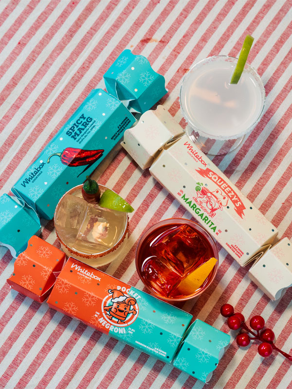 Whitebox Cocktails Christmas Crackers including the blue Spicy Margarita, the white Squeezy's Margarita and the orange and blue Pocket Negroni variants. All three are on top of a white and red pinstripe table.