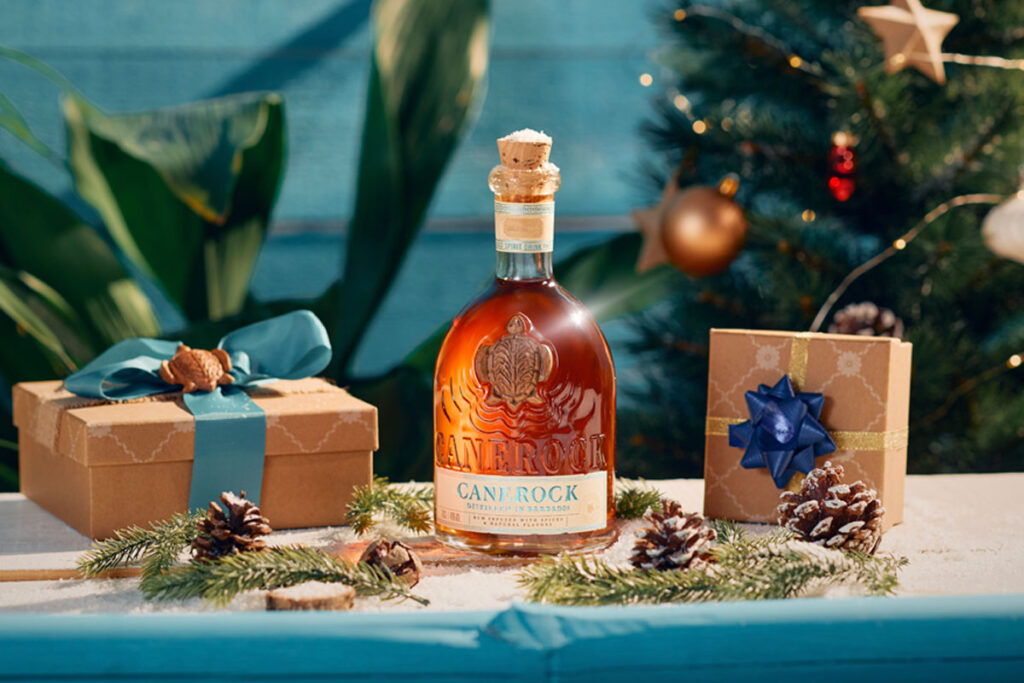 A bottle of Canerock Rum sits in the middle of a table filling with Christmas decorations and wrapped presents with a Christmas tree in the background.