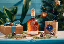 Canerock Rum brings the festive spirit to Sainsbury’s aisles A bottle of Canerock Rum sits in the middle of a table filling with Christmas decorations and wrapped presents with a Christmas tree in the background.