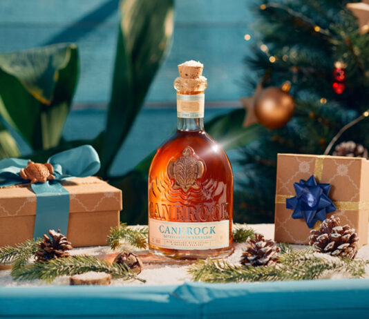 Canerock Rum brings the festive spirit to Sainsbury’s aisles A bottle of Canerock Rum sits in the middle of a table filling with Christmas decorations and wrapped presents with a Christmas tree in the background.