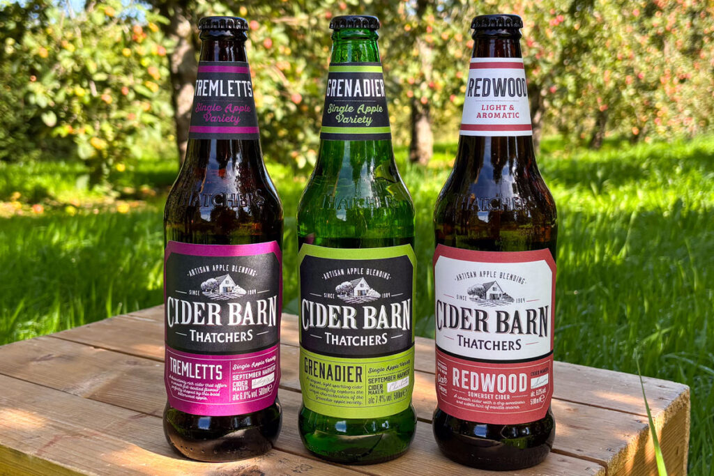 Thatchers Cider Barn bottles stand on a wooden crate in the middle of a grassy scene including Thatchers Redwood, Thatchers Grenadier and Thatchers Tremletts.