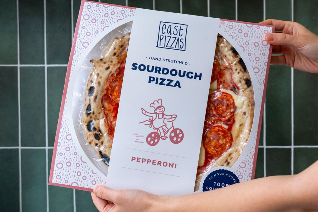 A person holds a pack of East Pizza Pepperoni Pizza.