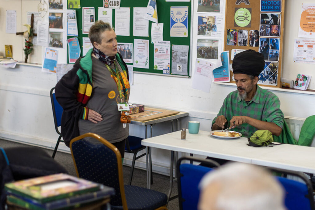 Two people are having a conversation; one is standing next to a table while the other is sitting at it eating a meal.