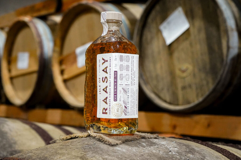 A bottle of Isle of Raasay whisky stands on top of a barrel in a warehouse with barrels in the background.