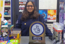 Allwyn shares the win with 20 National Lottery retailers A retailer stands in their store behind the counter with a National Lottery 'Millionaire Made Here' sign on the table.