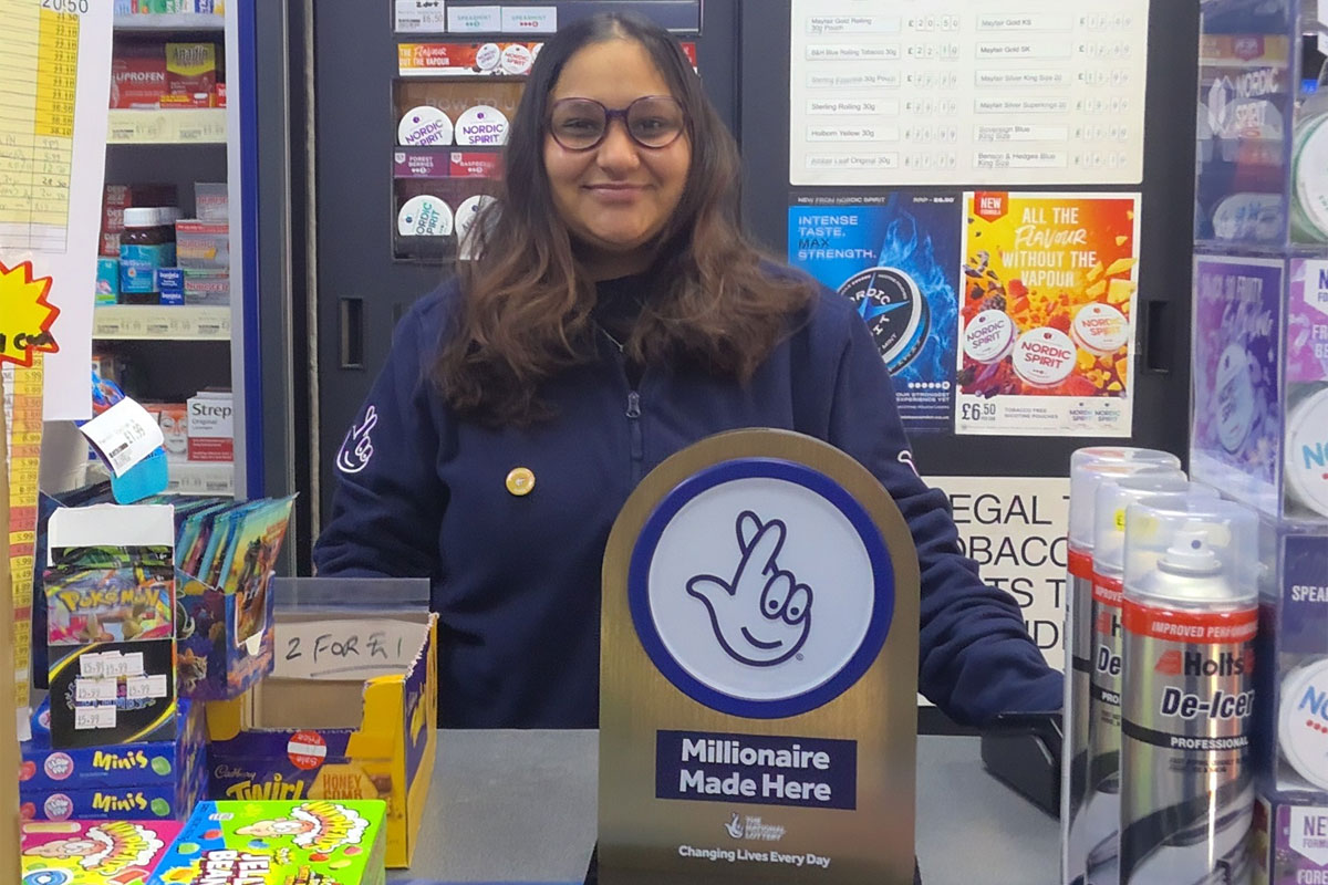 A retailer stands in their store behind the counter with a National Lottery 'Millionaire Made Here' sign on the table.