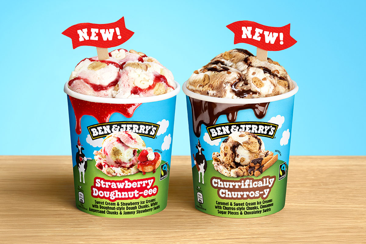 Tubs of Ben & Jerry's Strawberry Doughnut-eee and Churrifically Churros-y tubs stand on a wooden table against a light blue background.