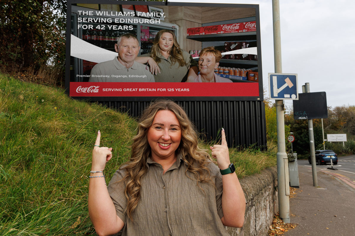 Sophie Williams, retailer at Premier Broadway Convenience Store, stands in front of a billboard as part of the 'The Bosses' campaign. The billboard includes an image of Sophie and her parents inside their store.