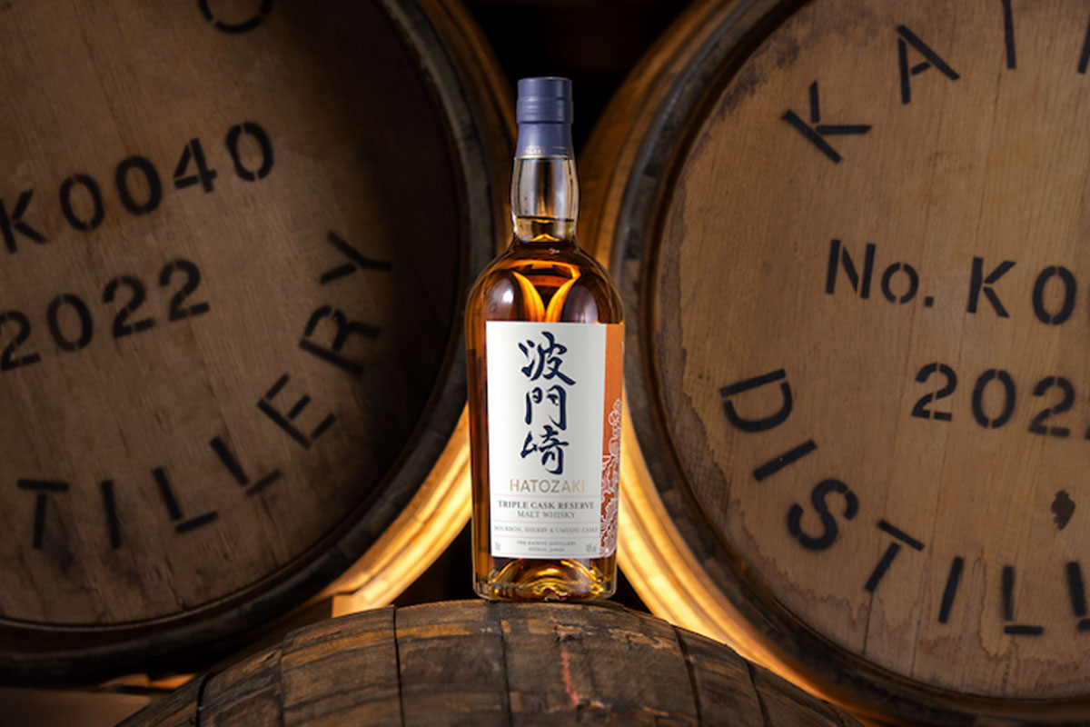 A bottle of Hatozaki Whisky Triple Cask Reserve is balanced on a wooden cask with wooden casks behind this.