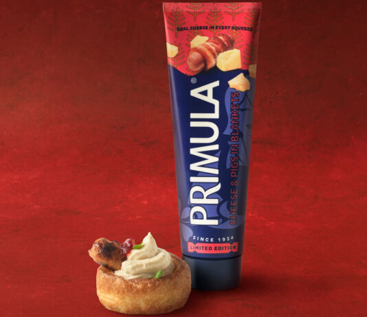 Primula brings back festive Pigs in Blanket flavour A tube of Primula Cheese Pigs in Blankets flavour stands next to a small bite serve using the cheese. This is against a red velvet background.