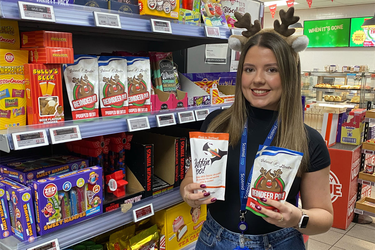 A woman wearing Reindeer antlers holds a pack of Puffin Poo and Reindeer Poo chocolates standing inside a Scotmid store.