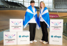 Allwyn announced as partner for Glasgow Commonwealth Games Two people stand in the Sir Chris Hoy Velodrome with Scotland flags draped across their shoulders with decorative boxes that have the Glasgow 2026 Commonwealth Games and Allwyn logos on them.