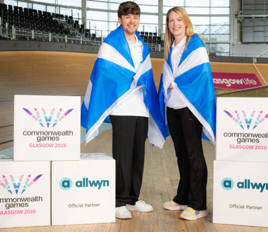 Allwyn announced as partner for Glasgow Commonwealth Games Two people stand in the Sir Chris Hoy Velodrome with Scotland flags draped across their shoulders with decorative boxes that have the Glasgow 2026 Commonwealth Games and Allwyn logos on them.
