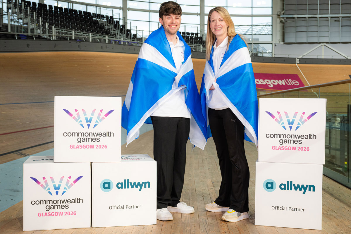 Two people stand in the Sir Chris Hoy Velodrome with Scotland flags draped across their shoulders with decorative boxes that have the Glasgow 2026 Commonwealth Games and Allwyn logos on them.