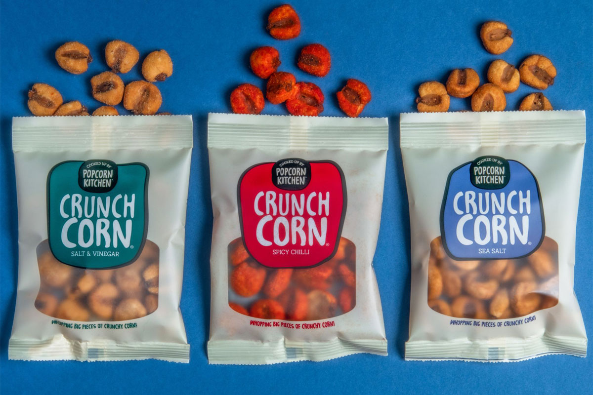 Pack of Popcorn Kitchen Crunch Corn lay open on a blue background with pieces of the corn spilled out from the top of the packs.