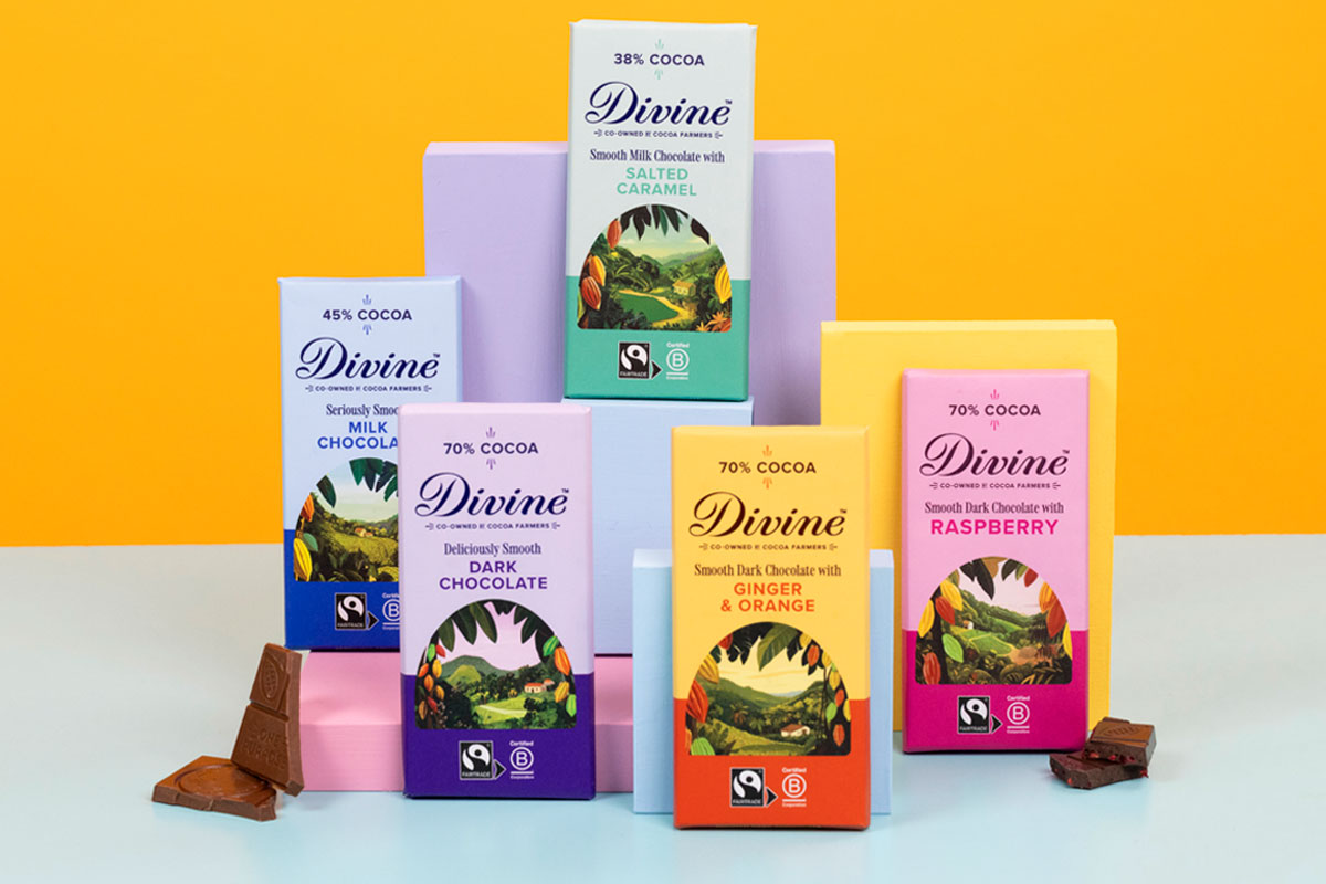 Bars of Divine Chocolate with the 2026 redesign are stacked against boxes to showcase the new looks. There are pieces of chocolate next to the bars and the background is an orange colour with a blue foreground.