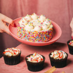 Finsbury launches first-to-market cake bento boxes A person places a large celebration cake on a pink plate down onto a table that already has four cupcakes sitting on it.