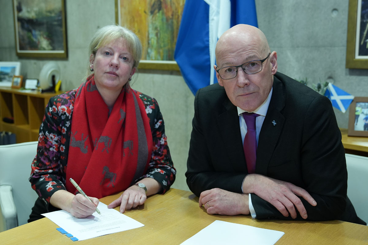 Shona Robison, finance secretary, and John Swinney, first minister, sit at a desk together with paper on the table and a saltire flag in the background.