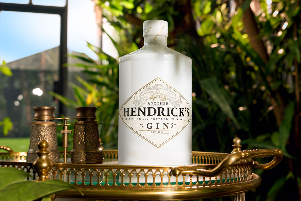 A bottle of Another Hendrick's Gin sits on a golden tabletop in a glass greenhouse with greenery around it.